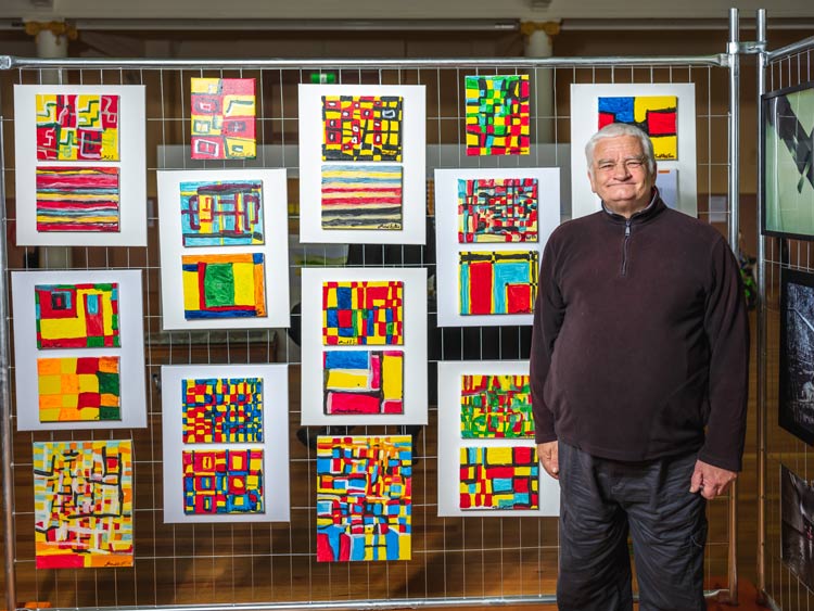 A man standing in front of a wire fence at City Hall with his coloured canvas art work hanging in the background