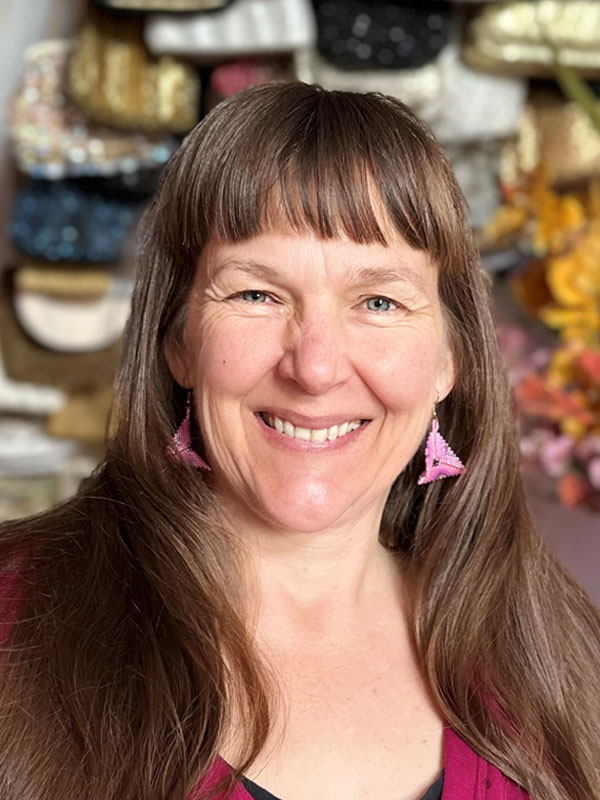 Lorenz Cherry – a woman’s head with long brown hair and laughing eyes and mouth. She is wearing triangular pink beaded earrings, with a colourful textured bag/purse artwork in the background. 